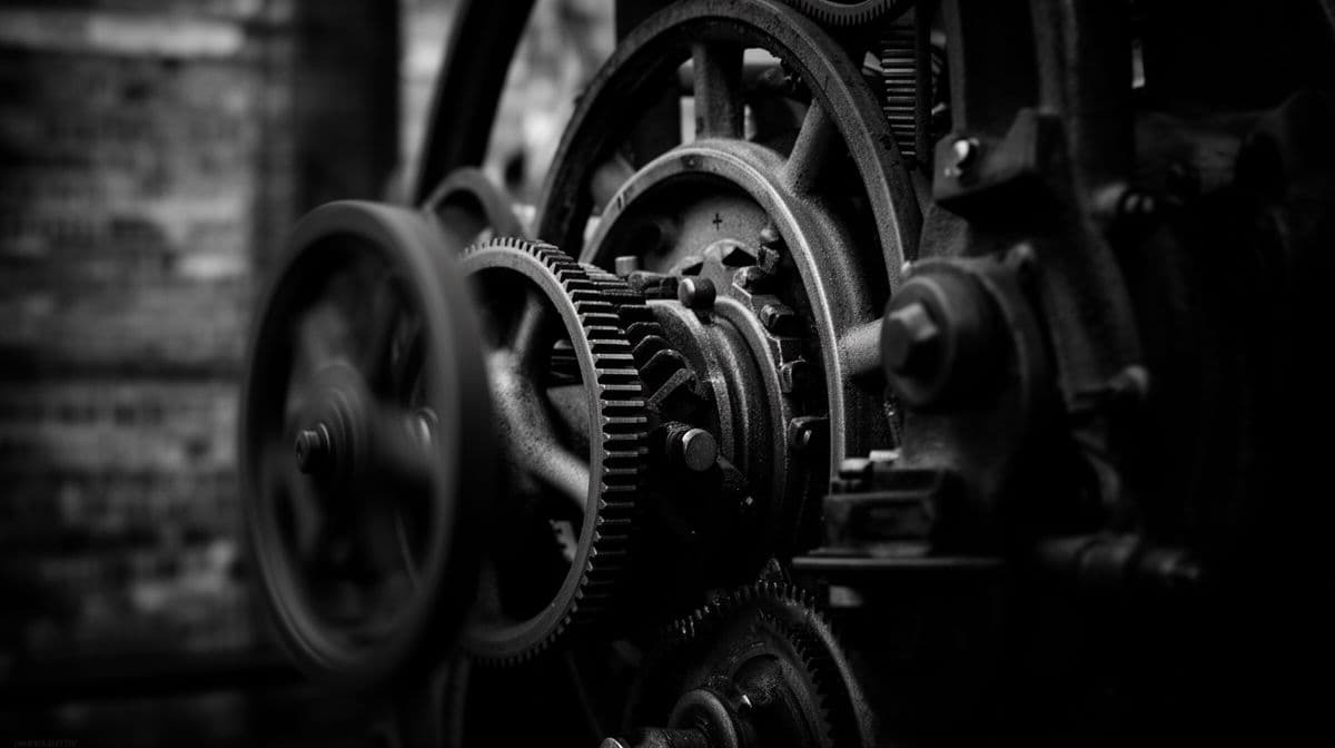 Prime plano em preto e branco de engrenagens industriais interligadas, com foco no mecanismo de transmissão de força, sugerindo complexidade e fluxo.